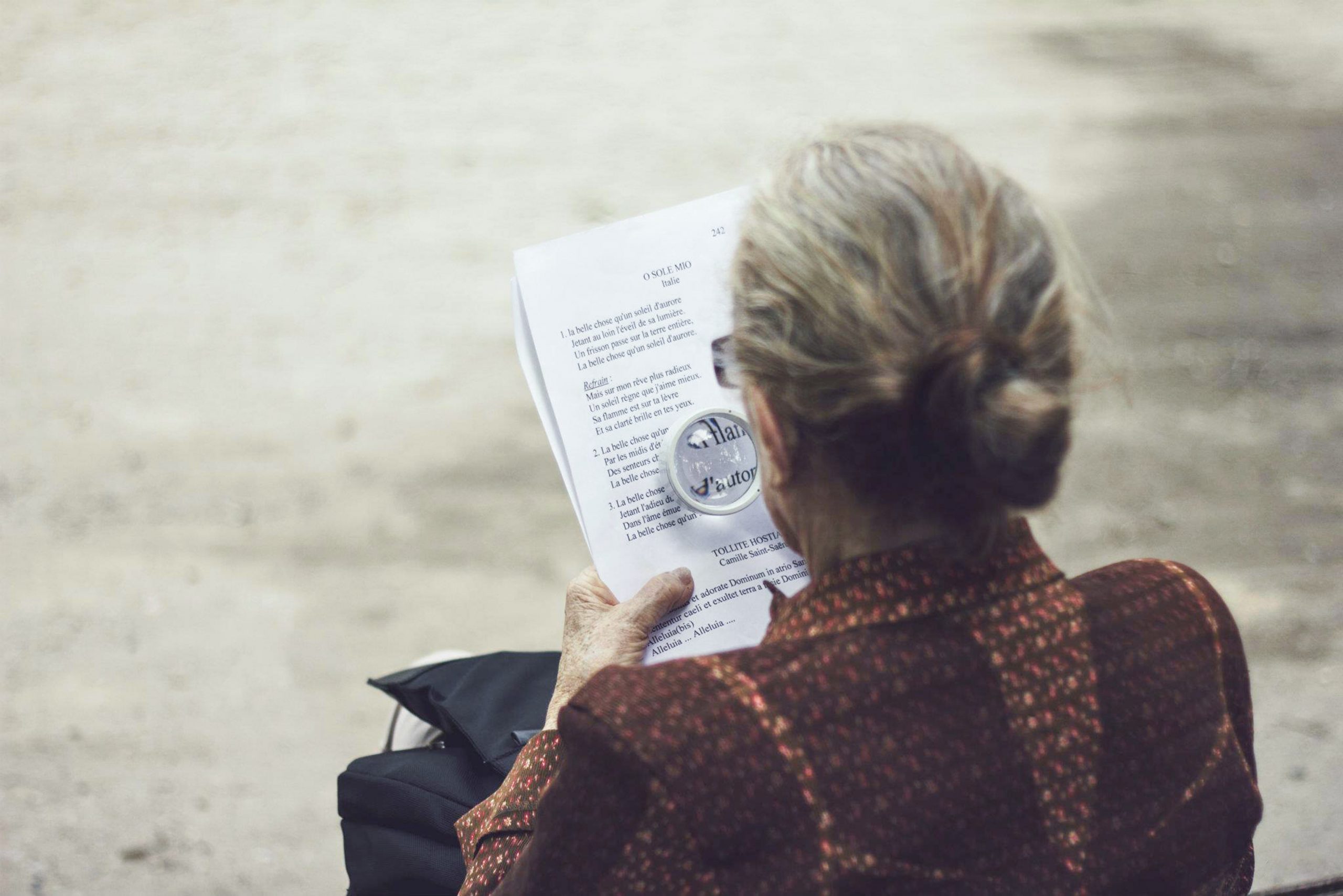 MCI - Was ist das? eine Definition der neurodegenerativen Erkrankung. Auf dem Bild zu sehen ist eine ältere Dame, die mit einer Leselupe ein Gedicht liest.